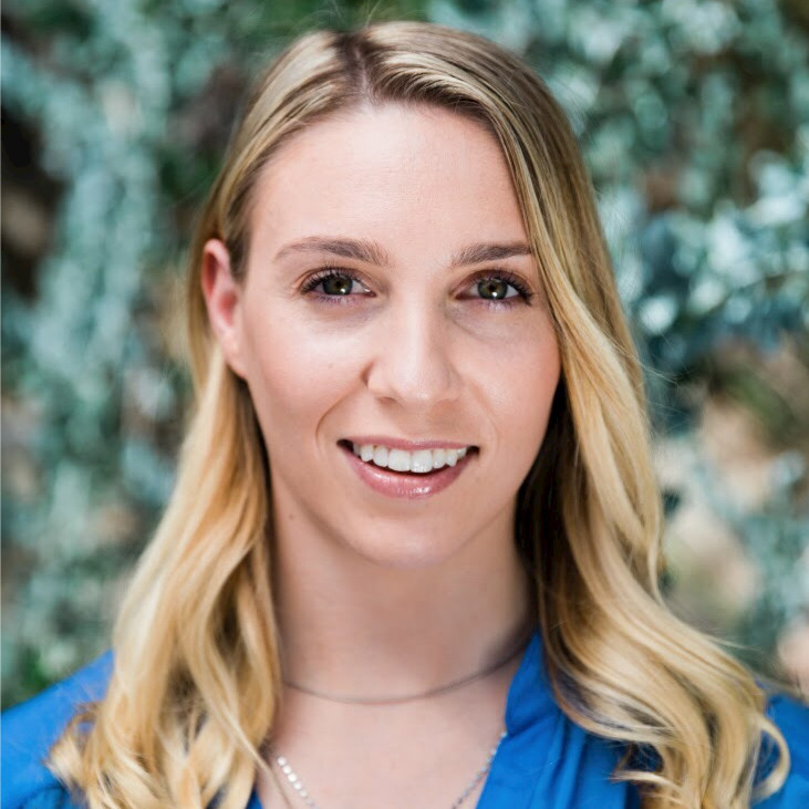 Headshot of a lady with long hair wearing blue top against a green leafy background. 